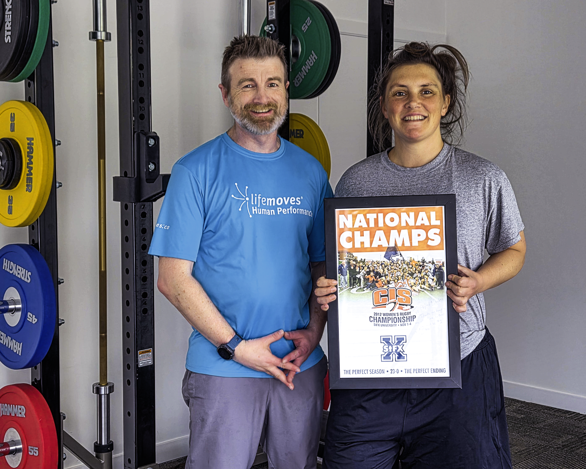 FST Client Beth Rugby 2026 Burnaby Alfred Ball, Level 3 FST practitioner (L), with retired athlete client Beth McNeil in 2026. She is holding a signed poster of ther CIS Women's Rugby Team Championship team from 2012. They are standing in front of a squat rack in Burnaby.