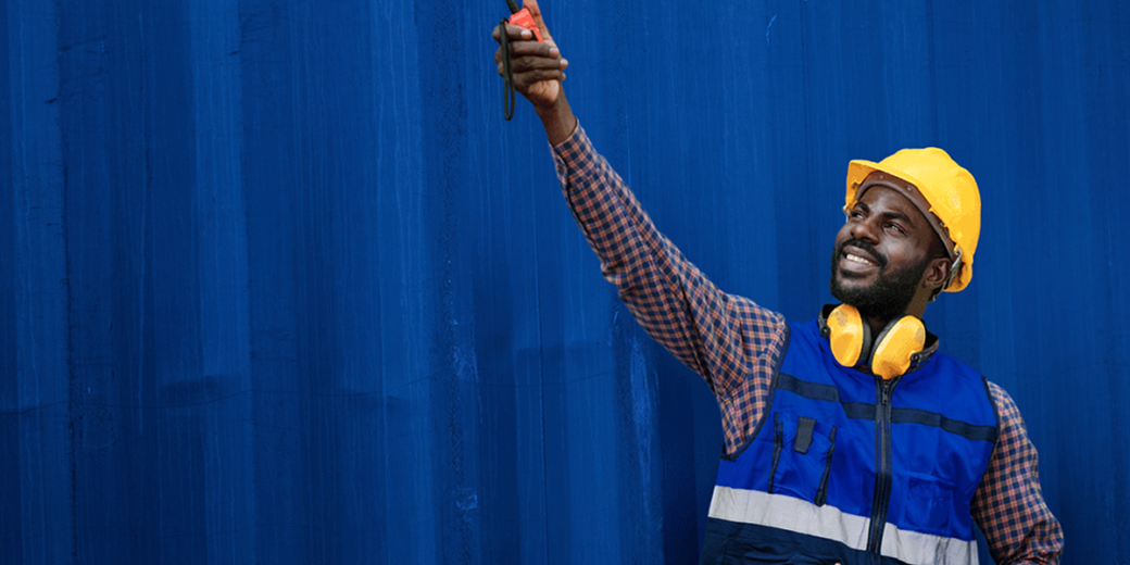 A smiling Black Male Longshoreman because he has returned to work after an injury. Wearing a yellow hard hat, blue safety vest, and yellow hearing protectors around his neck. He is pointing up with his right arm extended, holding a walkie-talkie. A dark blue crate is the background.